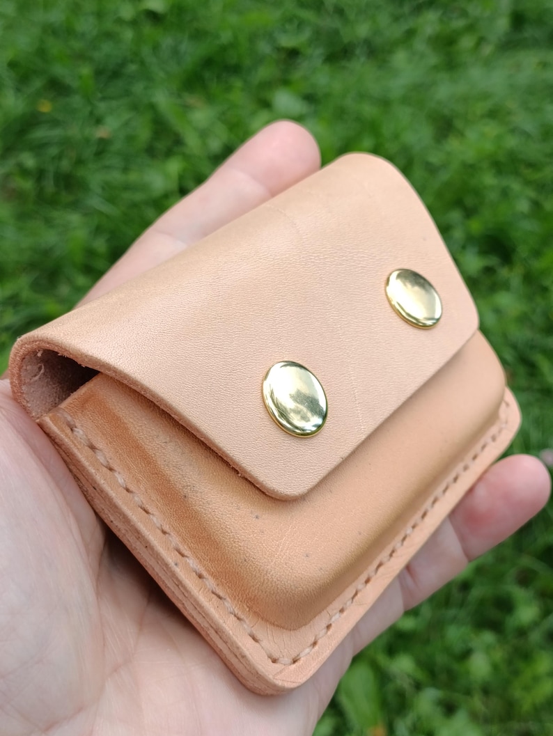 May include: A light tan leather pouch featuring a fold-over flap secured by two polished gold-tone metal snap buttons. The rectangular accessory is constructed with visible stitching along the edges and has a smooth, natural finish throughout.