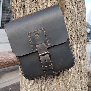 Pu&ograve; includere: Una borsa a tracolla in pelle marrone scuro con chiusura a fibbia e tracolla. La borsa &egrave; appesa a un tronco d'albero.