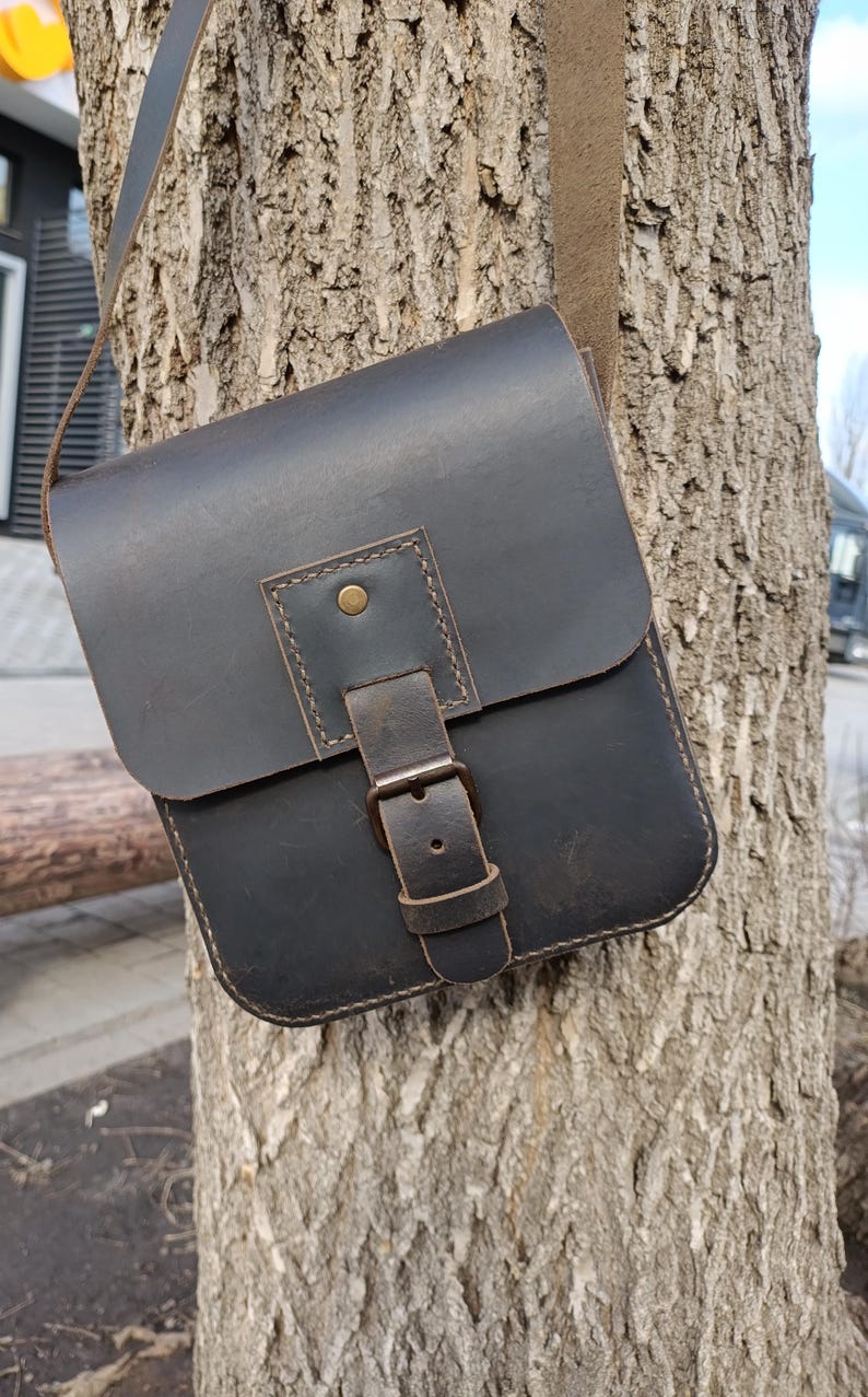 Pu&ograve; includere: Una borsa a tracolla in pelle marrone scuro con chiusura a fibbia e tracolla. La borsa &egrave; appesa a un tronco d'albero.