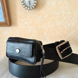 Puede incluir: Un cintur&oacute;n de cuero negro con un bolso rectangular sujeto a la correa. El bolso tiene una solapa con un bot&oacute;n de presi&oacute;n plateado y una cremallera lateral. La correa del cintur&oacute;n es lisa con una hebilla de metal plateado y varios agujeros.
