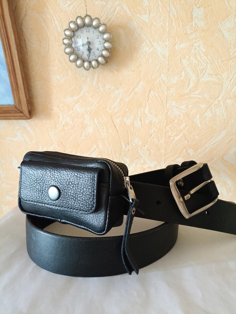 Puede incluir: Un cintur&oacute;n de cuero negro con un bolso rectangular sujeto a la correa. El bolso tiene una solapa con un bot&oacute;n de presi&oacute;n plateado y una cremallera lateral. La correa del cintur&oacute;n es lisa con una hebilla de metal plateado y varios agujeros.