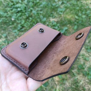 Puede incluir: Un tarjetero de cuero marr&oacute;n con cierre de solapa que cuenta con dos botones de presi&oacute;n de lat&oacute;n antiguo. El interior muestra un acabado de grano liso en el bolsillo y una superficie texturizada en la solapa, con costuras visibles.