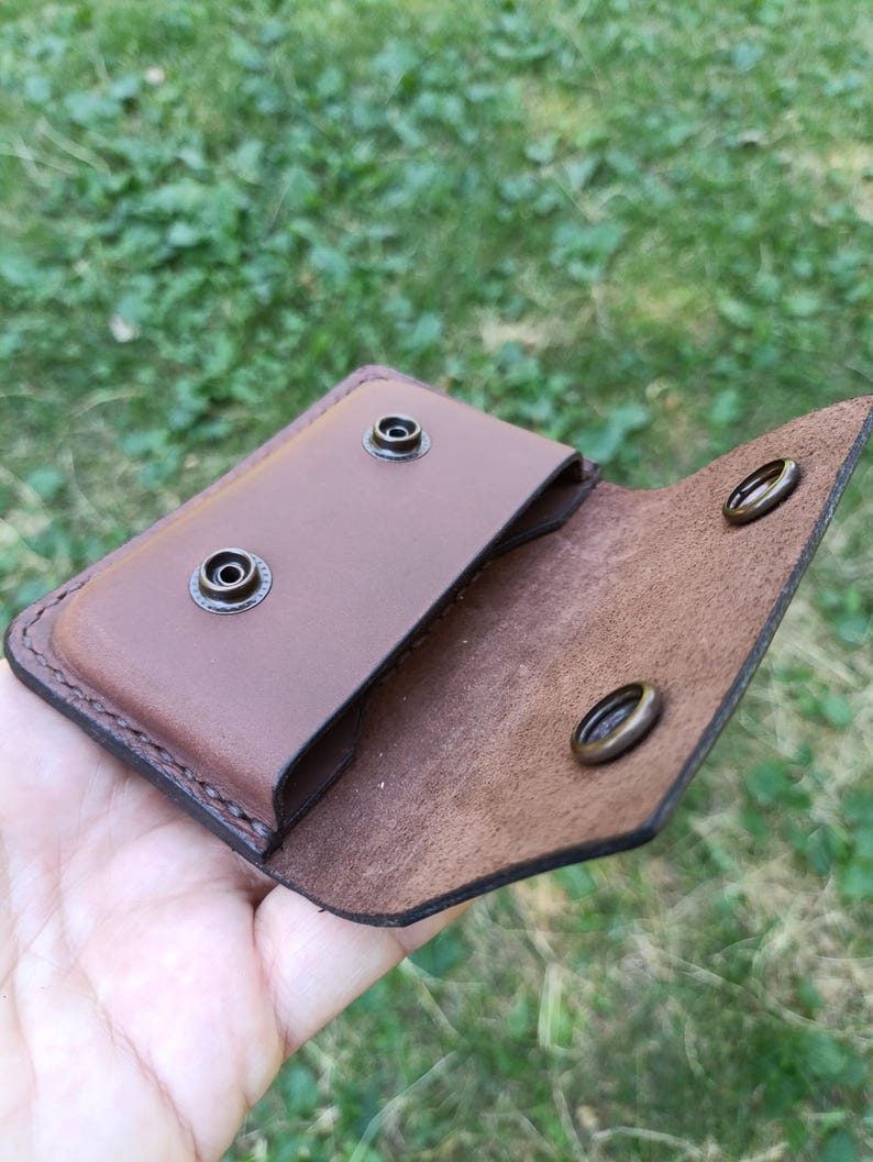 Puede incluir: Un tarjetero de cuero marr&oacute;n con cierre de solapa que cuenta con dos botones de presi&oacute;n de lat&oacute;n antiguo. El interior muestra un acabado de grano liso en el bolsillo y una superficie texturizada en la solapa, con costuras visibles.
