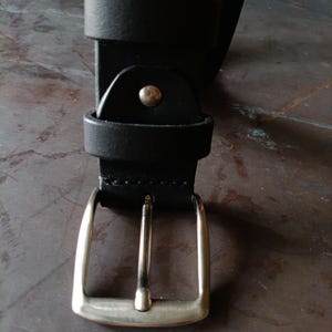 Puede incluir: Un cintur&oacute;n negro con una hebilla de metal plateado de una sola p&uacute;a. La correa del cintur&oacute;n est&aacute; asegurada con una trabilla de cuero negro a juego y un remache de metal plateado situado justo encima del conjunto de la hebilla.