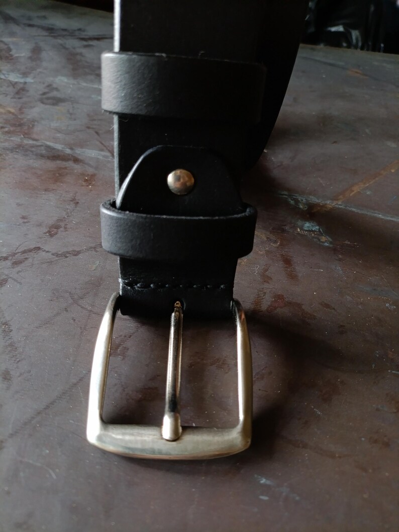 Puede incluir: Un cintur&oacute;n negro con una hebilla de metal plateado de una sola p&uacute;a. La correa del cintur&oacute;n est&aacute; asegurada con una trabilla de cuero negro a juego y un remache de metal plateado situado justo encima del conjunto de la hebilla.