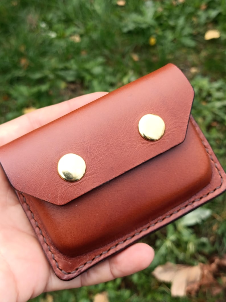 Puede incluir: Un tarjetero rectangular de cuero marr&oacute;n con cierre de solapa asegurado por dos botones de presi&oacute;n de metal dorado pulido. Los bordes presentan costuras visibles a lo largo del per&iacute;metro de la estructura de la cartera.