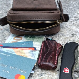 Peut inclure: Une pochette en cuir marron avec une fermeture &agrave; pression en laiton et une fermeture &eacute;clair, une pile de cartes bancaires, un &eacute;tui &agrave; briquet en cuir textur&eacute; rouge fonc&eacute; avec un briquet Cricket et un outil multifonction noir.