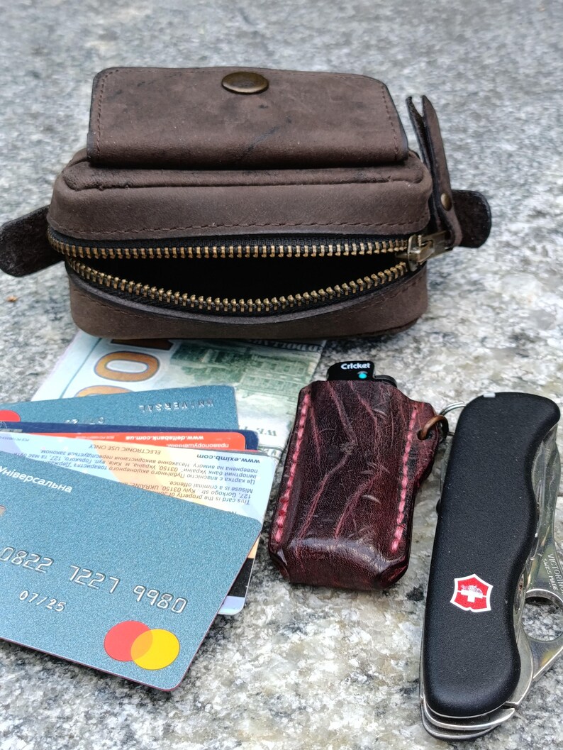 Peut inclure: Une pochette en cuir marron avec une fermeture &agrave; pression en laiton et une fermeture &eacute;clair, une pile de cartes bancaires, un &eacute;tui &agrave; briquet en cuir textur&eacute; rouge fonc&eacute; avec un briquet Cricket et un outil multifonction noir.