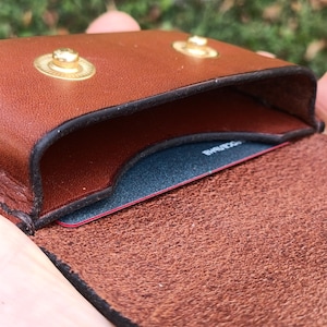 May include: A brown leather card holder featuring two gold-toned metal snap buttons on the top flap. The interior compartment contains a dark blue card with visible text, and the leather has a smooth exterior finish with a textured inner side.
