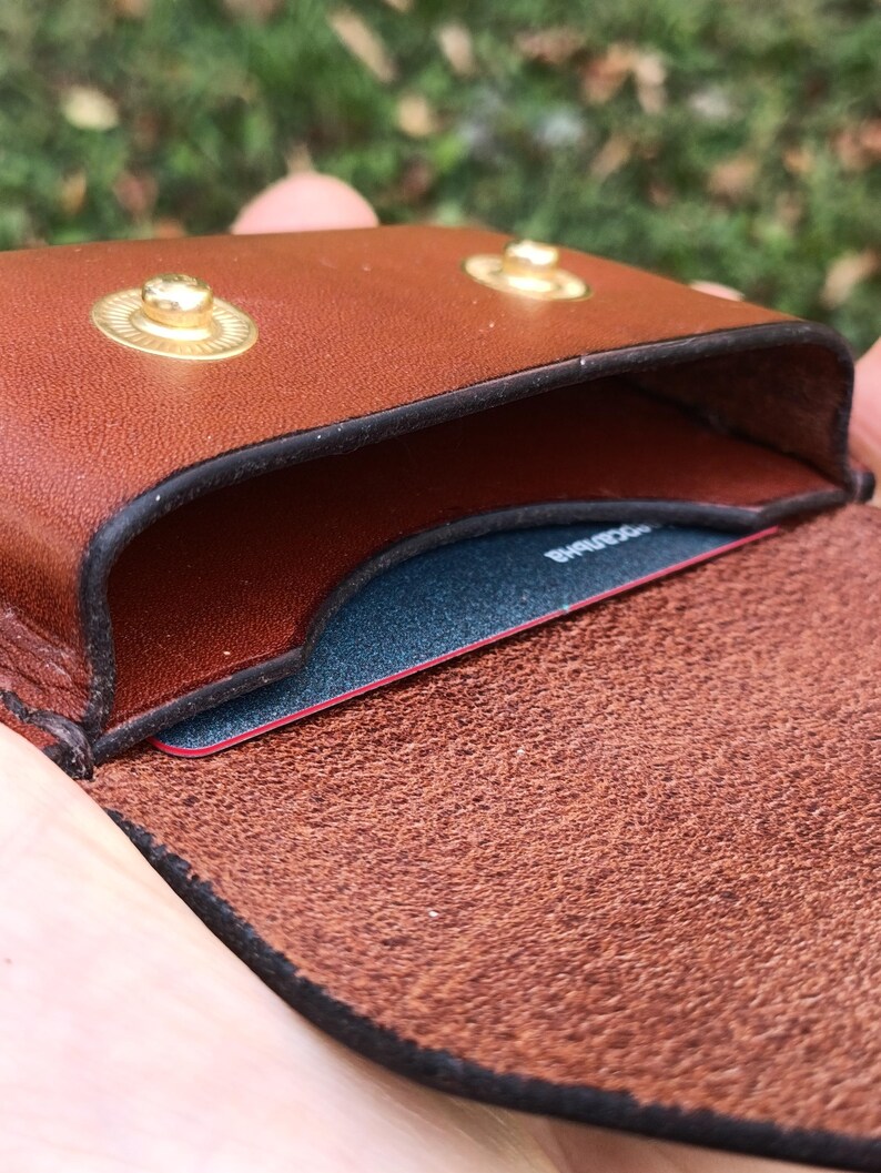 May include: A brown leather card holder featuring two gold-toned metal snap buttons on the top flap. The interior compartment contains a dark blue card with visible text, and the leather has a smooth exterior finish with a textured inner side.