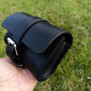 May include: A black leather roll-up watch case with a flap closure and a buckle. The case is cylindrical and has a strap that wraps around it. The background is blurred green grass.