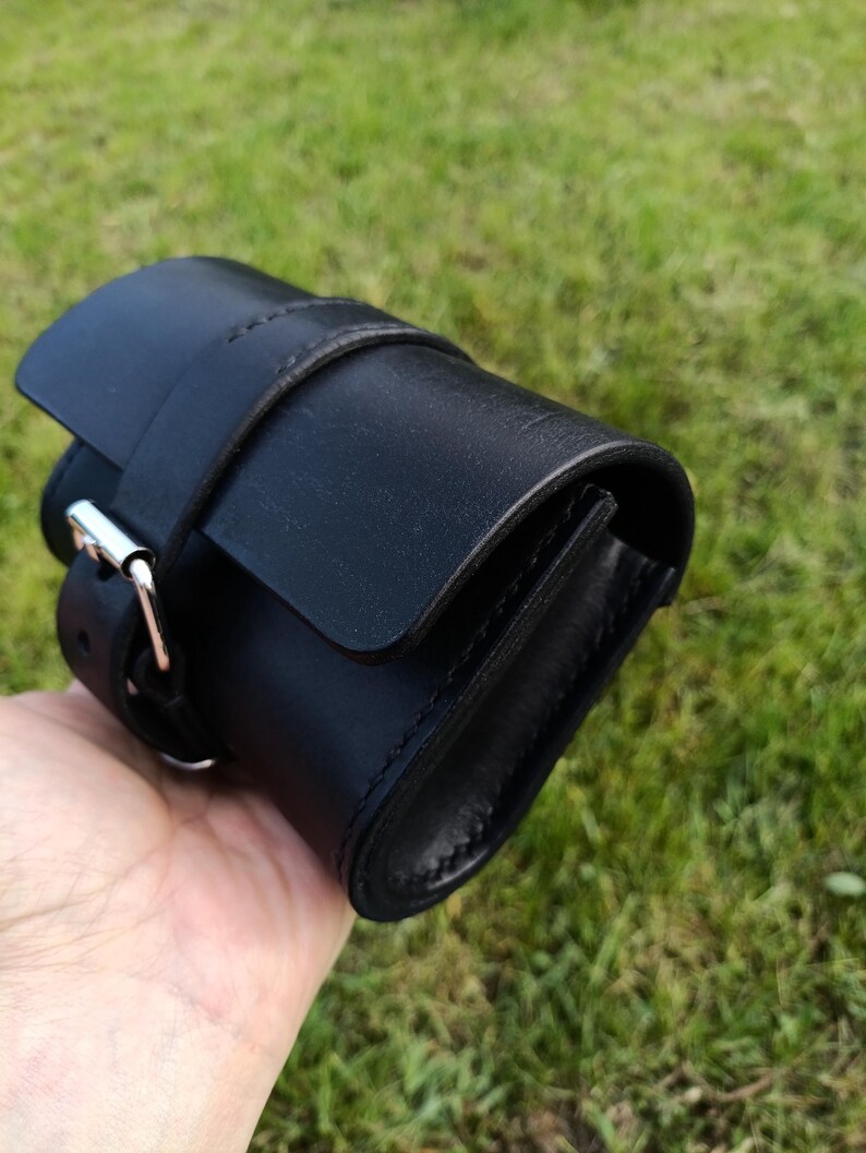 May include: A black leather roll-up watch case with a flap closure and a buckle. The case is cylindrical and has a strap that wraps around it. The background is blurred green grass.