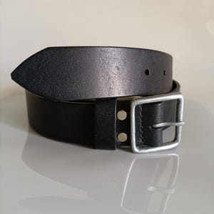 May include: A black leather belt with a silver-colored square buckle. The belt has a pointed end and two silver rivets near the buckle. The leather appears to have a slightly worn texture.