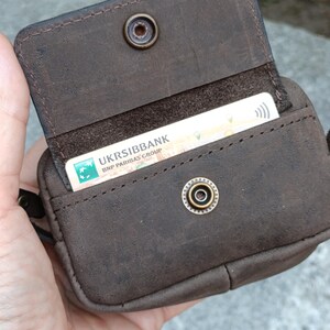 Peut inclure: Un porte-cartes en cuir marron fonc&eacute; avec une fermeture &agrave; rabat s&eacute;curis&eacute;e par un bouton-pression en m&eacute;tal. L'int&eacute;rieur contient une carte de paiement blanche avec le texte UKRSIBBANK BNP PARIBAS GROUP visible. La surface a un fini mat textur&eacute;.