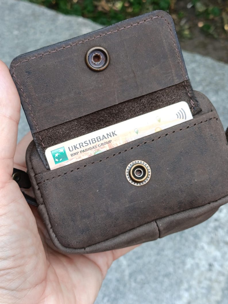 Peut inclure: Un porte-cartes en cuir marron fonc&eacute; avec une fermeture &agrave; rabat s&eacute;curis&eacute;e par un bouton-pression en m&eacute;tal. L'int&eacute;rieur contient une carte de paiement blanche avec le texte UKRSIBBANK BNP PARIBAS GROUP visible. La surface a un fini mat textur&eacute;.