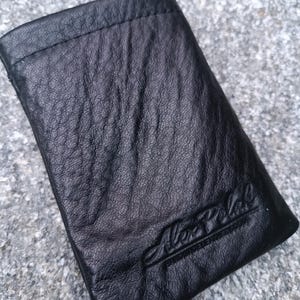 May include: A black leather pouch with a textured, wrinkled surface and a stitched top edge. The name Alex Polak is embossed in a cursive script in the bottom right corner of the rectangular accessory, which features a simple, flat design.