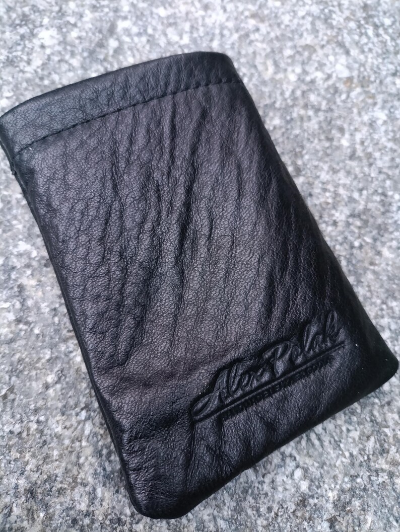 May include: A black leather pouch with a textured, wrinkled surface and a stitched top edge. The name Alex Polak is embossed in a cursive script in the bottom right corner of the rectangular accessory, which features a simple, flat design.