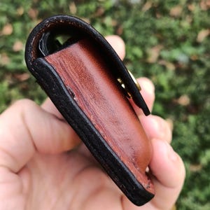 May include: A small leather pouch featuring a smooth, reddish-brown main body with thick, dark-brown burnished edges. The pouch is secured with a gold-toned metal snap fastener visible on the side, showcasing detailed stitching and layered construction.