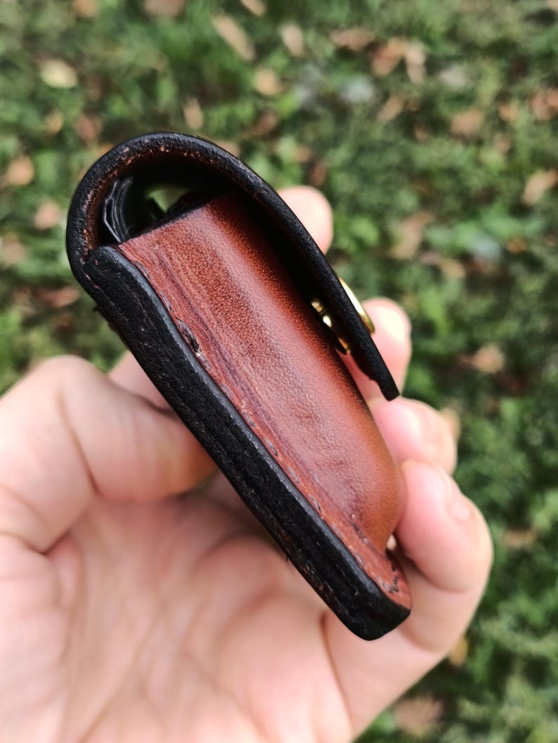 May include: A small leather pouch featuring a smooth, reddish-brown main body with thick, dark-brown burnished edges. The pouch is secured with a gold-toned metal snap fastener visible on the side, showcasing detailed stitching and layered construction.