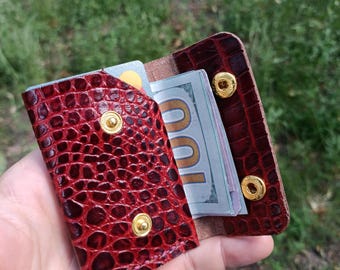 Puede incluir: Una cartera pequeña y rectangular de color rojo oscuro con un patrón texturizado de piel de cocodrilo. La cartera cuenta con botones de presión de metal dorado y contiene un billete de cien dólares doblado y una tarjeta de crédito.