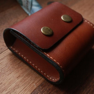 May include: A brown leather case with a flap closure and two brass snaps. The case is stitched with white thread and has a dark brown lining.