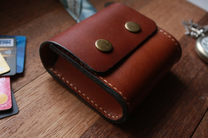 May include: A brown leather case with a flap closure and two brass snaps. The case is stitched with white thread and has a dark brown lining.