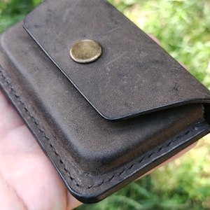 May include: A dark brown leather card holder with a matte finish and visible stitching along the edges. The rectangular accessory features a single flap closure secured by a circular, antique brass-toned metal snap button centered on the front.