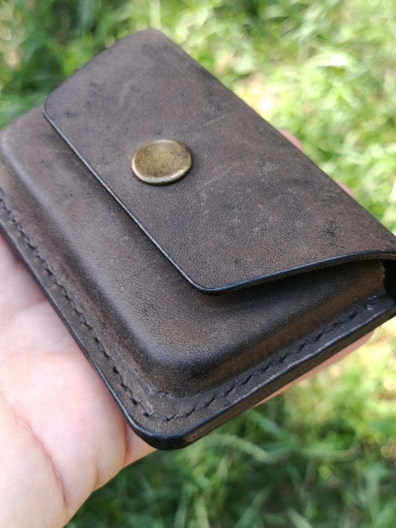 May include: A dark brown leather card holder with a matte finish and visible stitching along the edges. The rectangular accessory features a single flap closure secured by a circular, antique brass-toned metal snap button centered on the front.