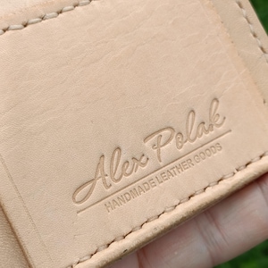 May include: A light tan leather wallet featuring visible cream-colored stitching along the edges. The surface is embossed with the text Alex Polak and the words HANDMADE LEATHER GOODS positioned directly underneath the name in a clean font.