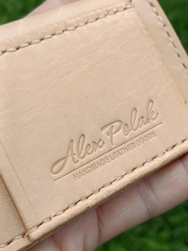 May include: A light tan leather wallet featuring visible cream-colored stitching along the edges. The surface is embossed with the text Alex Polak and the words HANDMADE LEATHER GOODS positioned directly underneath the name in a clean font.