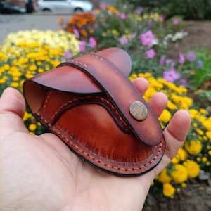 K&ouml;nnte beinhalten: Eine handgefertigte braune Lederh&uuml;lle mit geschwungenem Design, gesichert durch einen Riemen und einen Messingknopf. Die H&uuml;lle hat sichtbare N&auml;hte an den Kanten und wird in einer Hand gehalten, mit einem verschwommenen floralen Hintergrund.