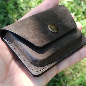 May include: A dark brown leather card holder with a rectangular shape and rounded corners. The accessory features a flap closure secured by a single circular brass-toned metal snap button. The edges are finished with visible dark stitching.