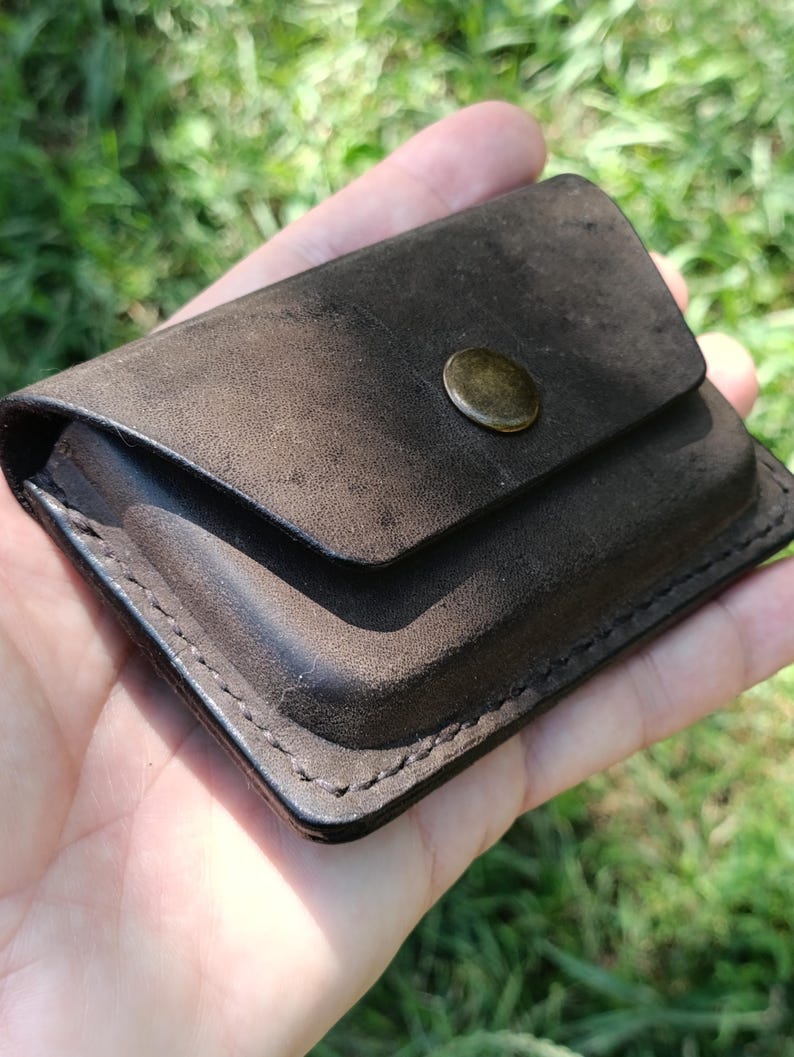 May include: A dark brown leather card holder with a rectangular shape and rounded corners. The accessory features a flap closure secured by a single circular brass-toned metal snap button. The edges are finished with visible dark stitching.