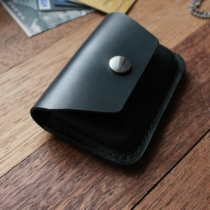 Puede incluir: Una cartera de cuero negro con cierre de presi&oacute;n y un bot&oacute;n plateado. La cartera est&aacute; sobre una superficie de madera.