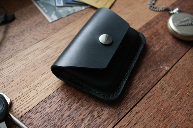 Puede incluir: Una cartera de cuero negro con cierre de presi&oacute;n y un bot&oacute;n plateado. La cartera est&aacute; sobre una superficie de madera.