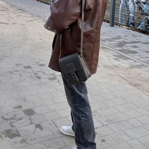 Pu&ograve; includere: Una persona indossa una giacca di pelle marrone e jeans blu scuro con una borsa a tracolla in pelle nera. La borsa ha una chiusura a fibbia e una tracolla che passa sopra la spalla.