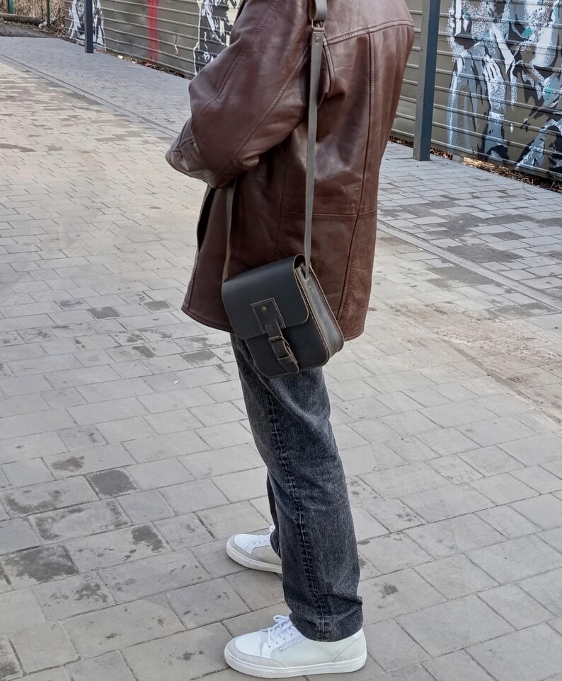 Pu&ograve; includere: Una persona indossa una giacca di pelle marrone e jeans blu scuro con una borsa a tracolla in pelle nera. La borsa ha una chiusura a fibbia e una tracolla che passa sopra la spalla.