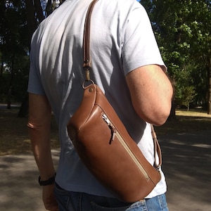 May include: A brown leather crossbody bag with a zippered pocket and a long adjustable strap. The bag is worn by a person in a casual setting.