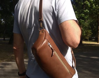 May include: A brown leather crossbody bag with a zippered pocket and a long adjustable strap. The bag is worn by a person in a casual setting.