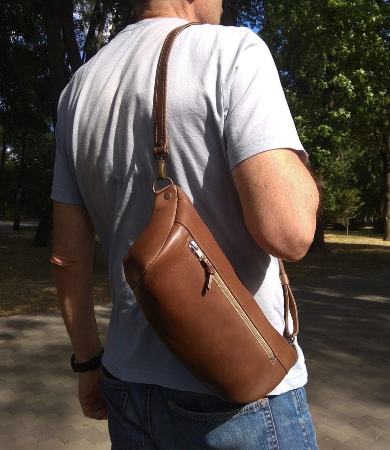 May include: A brown leather crossbody bag with a zippered pocket and a long adjustable strap. The bag is worn by a person in a casual setting.