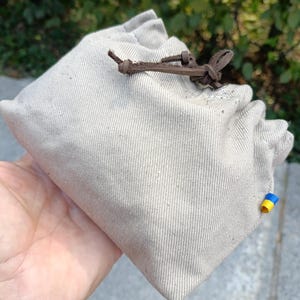 Peut inclure: Une pochette &agrave; cordon en toile beige avec une fermeture en cuir marron fonc&eacute;. Le tissu pr&eacute;sente une texture tiss&eacute;e visible et une petite &eacute;tiquette rectangulaire &agrave; rayures horizontales bleues et jaunes est fix&eacute;e sur la couture lat&eacute;rale.
