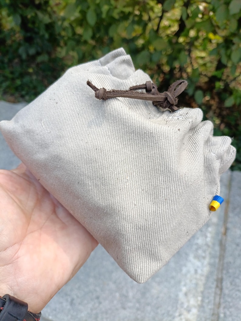 Peut inclure: Une pochette &agrave; cordon en toile beige avec une fermeture en cuir marron fonc&eacute;. Le tissu pr&eacute;sente une texture tiss&eacute;e visible et une petite &eacute;tiquette rectangulaire &agrave; rayures horizontales bleues et jaunes est fix&eacute;e sur la couture lat&eacute;rale.