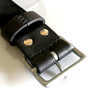 以下が含まれることがあります： シルバー色のバックルと2つのシルバーリベットが付いた黒い革ベルトのクローズアップ。ベルトはステッチされており、ベルトの端を保持するためのループが付いています。バックルは長方形です。