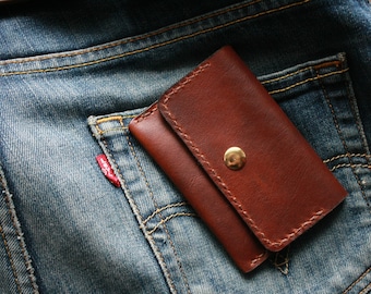 May include: A brown leather wallet with a gold snap closure rests in the back pocket of a pair of blue denim jeans. The wallet is rectangular and appears to be hand-stitched. The jeans have a red tag on the pocket.