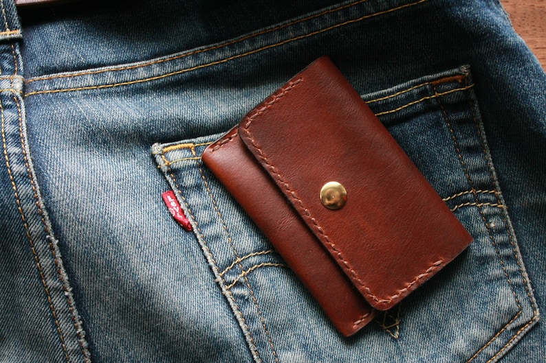 May include: A brown leather wallet with a gold snap closure rests in the back pocket of a pair of blue denim jeans. The wallet is rectangular and appears to be hand-stitched. The jeans have a red tag on the pocket.