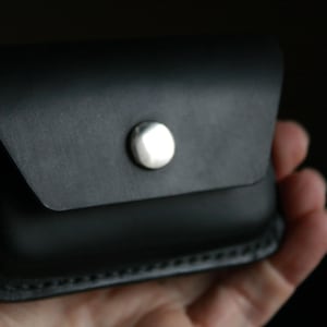 Puede incluir: Una cartera de cuero negro con cierre de presi&oacute;n plateado. La cartera est&aacute; doblada por la mitad y tiene un borde cosido.