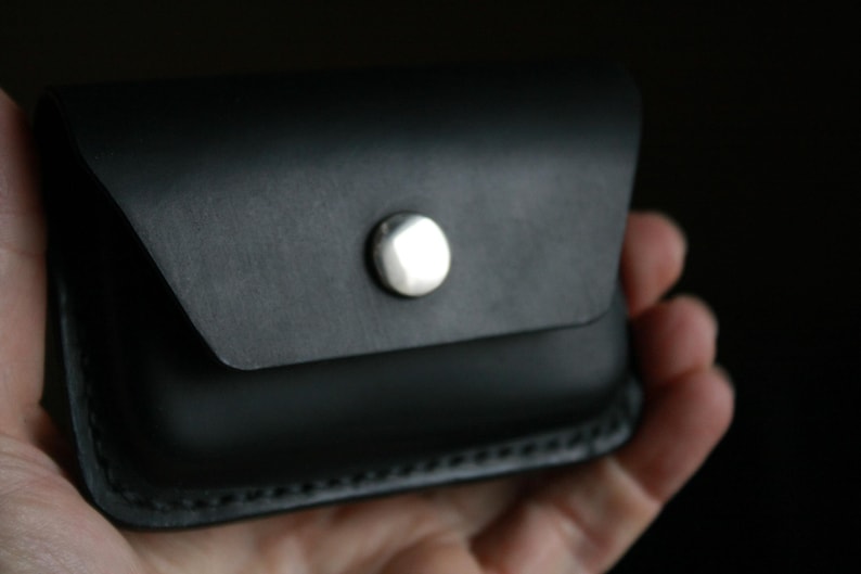Puede incluir: Una cartera de cuero negro con cierre de presi&oacute;n plateado. La cartera est&aacute; doblada por la mitad y tiene un borde cosido.