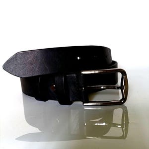 K&ouml;nnte beinhalten: Ein schwarzer Lederg&uuml;rtel mit einer silberfarbenen Schnalle. Der G&uuml;rtel besteht aus einem St&uuml;ck Leder und hat ein schlichtes, klassisches Design. Die rechteckige Schnalle hat eine polierte Oberfl&auml;che.