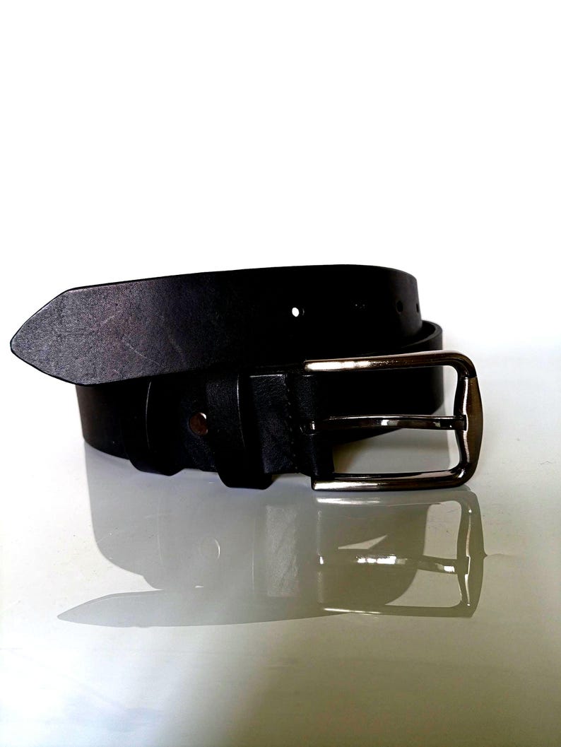 K&ouml;nnte beinhalten: Ein schwarzer Lederg&uuml;rtel mit einer silberfarbenen Schnalle. Der G&uuml;rtel besteht aus einem St&uuml;ck Leder und hat ein schlichtes, klassisches Design. Die rechteckige Schnalle hat eine polierte Oberfl&auml;che.