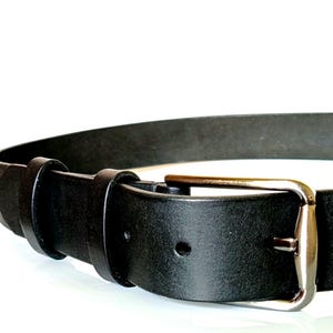 K&ouml;nnte beinhalten: Nahaufnahme eines schwarzen Lederg&uuml;rtels mit silberfarbener Schnalle. Der G&uuml;rtel hat ein schlichtes, klassisches Design mit einer einzelnen Schlaufe und einer rechteckigen Schnalle. Das Leder wirkt glatt und der G&uuml;rtel ist vor wei&szlig;em Hintergrund platziert.